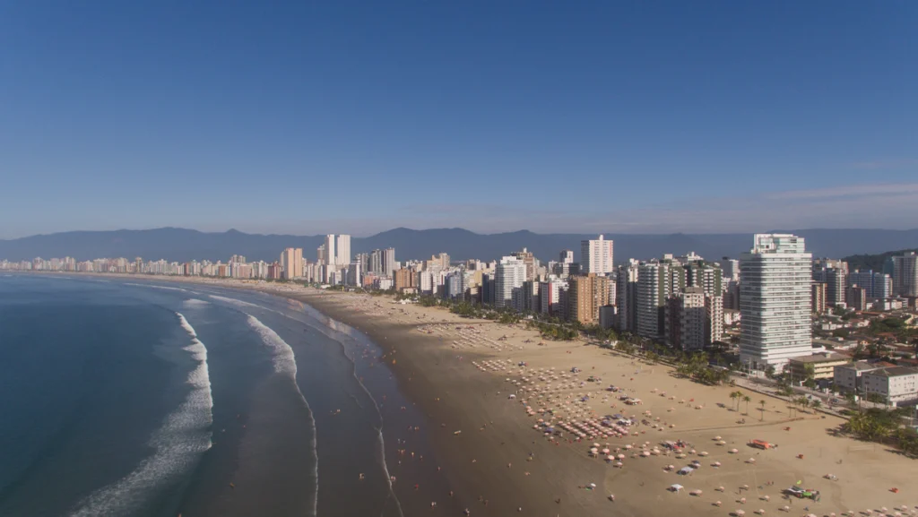 Orla de Praia Grande SP em um dia de sol, com alguns guarda-sóis na areia da praia. Vista aérea feita com drone no Canto do Forte.